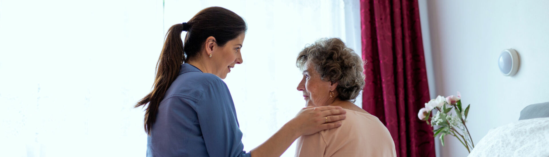 Home Caregiver Comforting Senior Woman Patient Sitting in Her Bedroom. Nurse Putting Her Hand Over Elderly Patient Shoulder in Hospital Room.