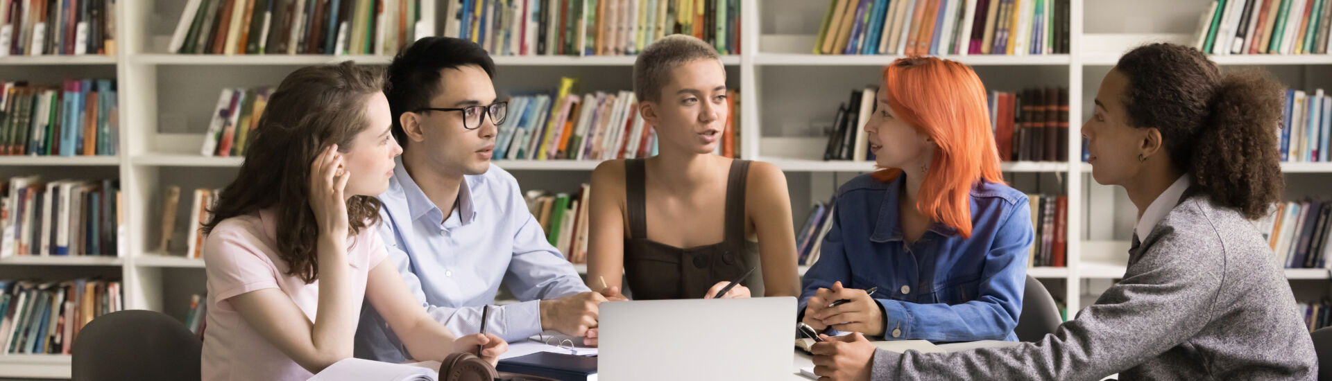 Multiethnic teenage students gather in library around table with laptop