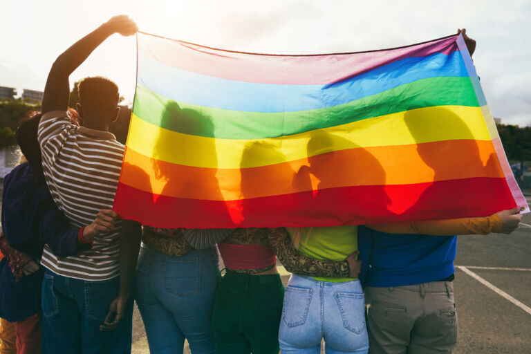 Young diverse people having fun holding lgbt rainbow flag outdoor – Main focus on african guy back