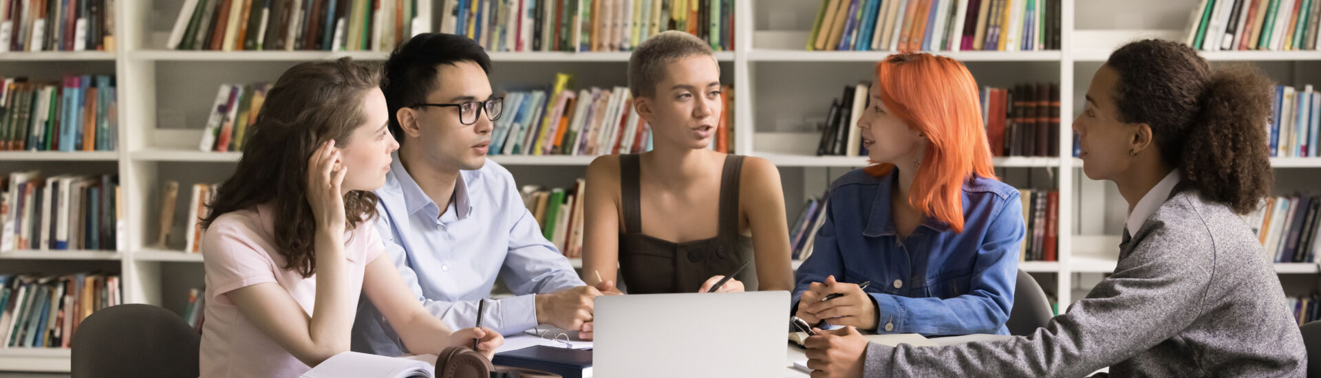 Multiethnic teenage students gather in library around table with laptop