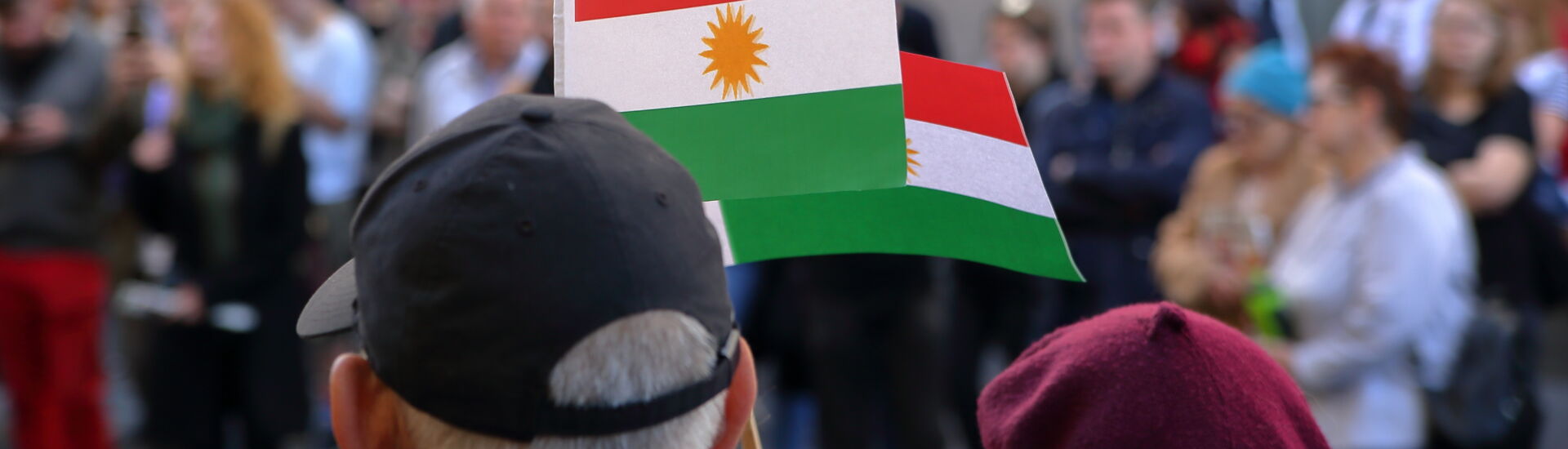 Two flags of Kurds hold by adult woman and men, standing on their back, during street protest against Turkish invasion in Syria, in background other protesters in soft focus