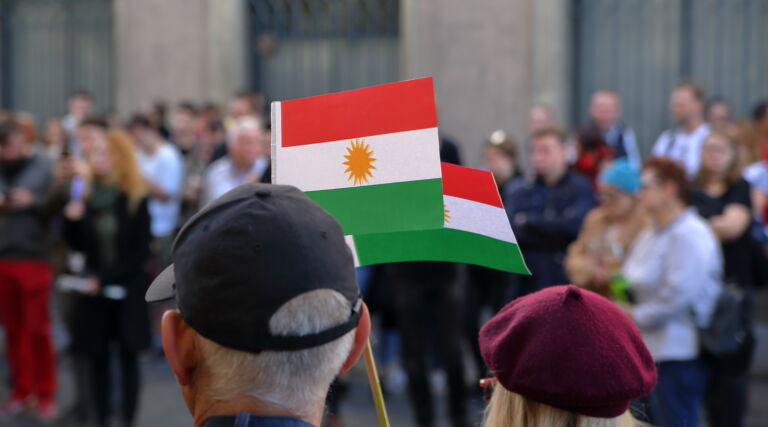 Two flags of Kurds hold by adult woman and men, standing on their back, during street protest against Turkish invasion in Syria, in background other protesters in soft focus