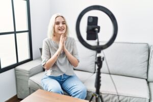 Young caucasian woman recording vlog tutorial with smartphone at home praying with hands together asking for forgiveness smiling confident.